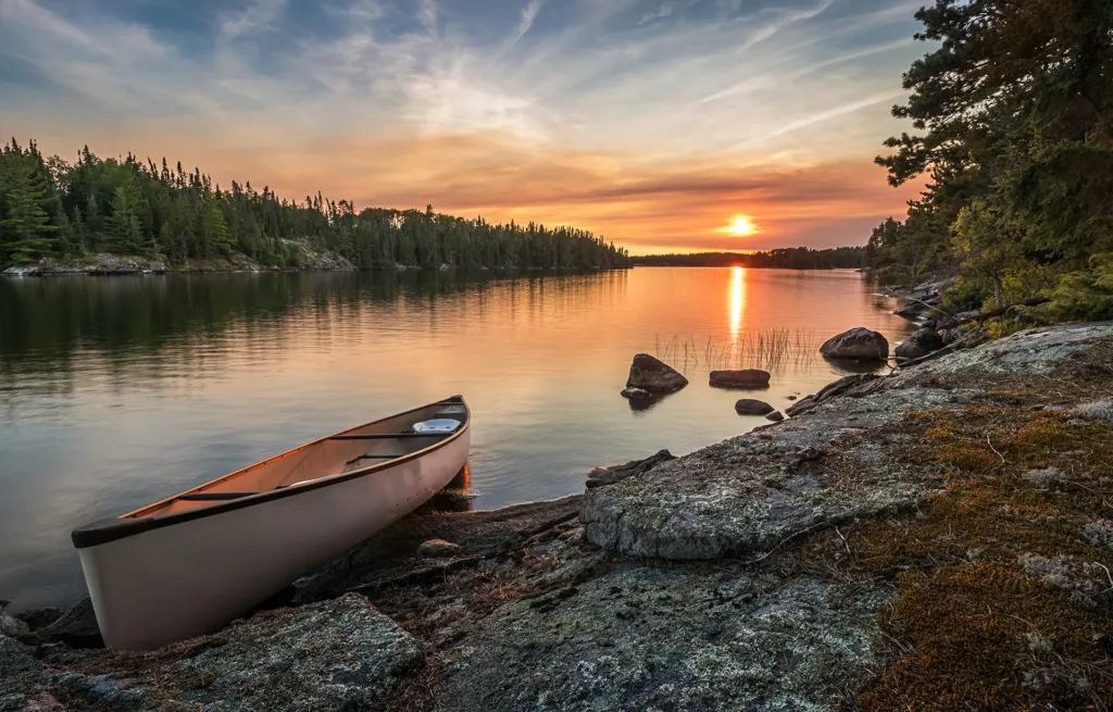 Canoe resting on a rocky lakeshore at sunset, with boreal forest lining the calm water