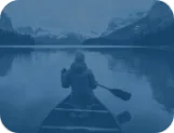 Person paddling a canoe across a calm mountain lake toward snow-capped peaks under low clouds