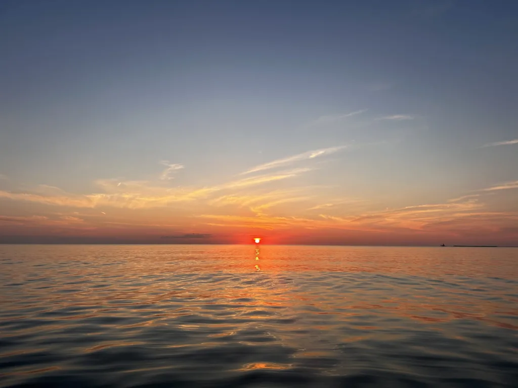 Sun setting on the horizon over calm open water, with a distant lighthouse on a breakwater