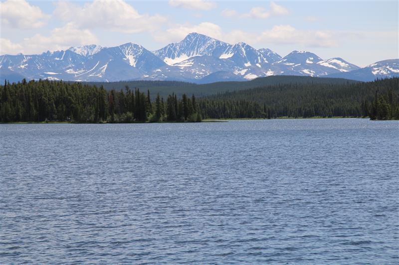 Lake with forested shoreline below snow-capped mountains on a clear summer day